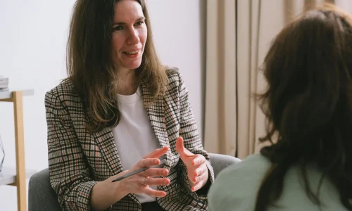 Pensive woman psychologist with brown hair in stylish clothes sitting and talking with unrecognizable female in light room